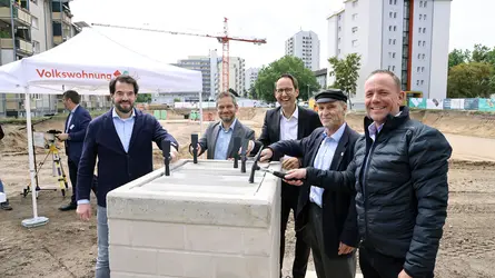 fünf Männer hauen mit einem schwarzen Hammer auf einen Grundstein und lächeln dabei