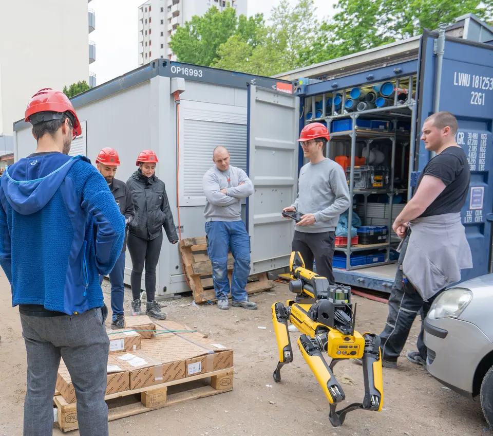 weisenburger Team mit Roboter vor einer Palette  Ein Boston Dynamics Spot Roboter vor einer Palette umgeben von vielen Mitarbeitern