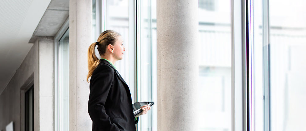 Eine Frau in einem Bürogebäude lässt den Blick aus Fenster in die Ferne schweifen