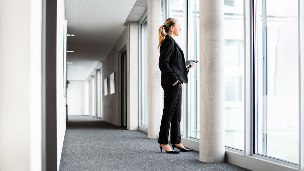 Eine Frau in einem Bürogebäude lässt den Blick aus Fenster in die Ferne schweifen