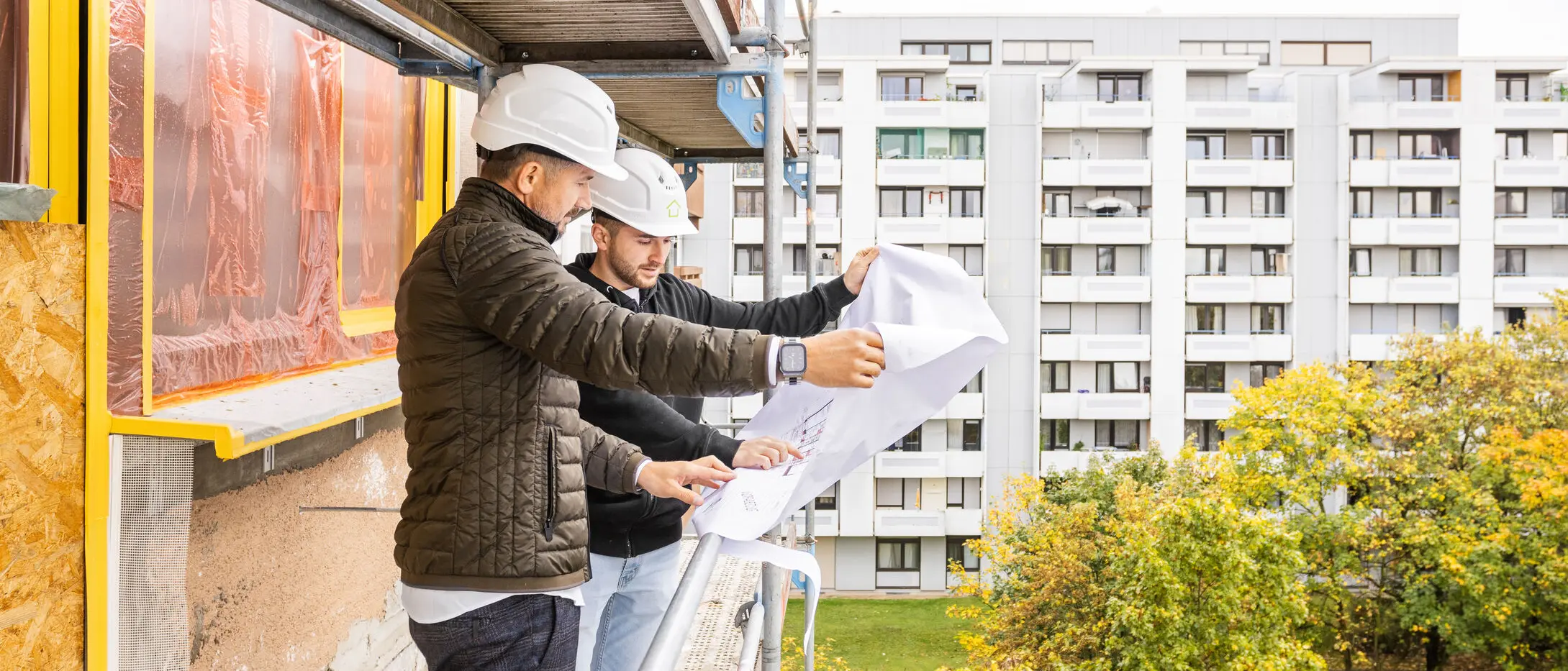 Zwei Männer mit Bauhelmen stehen auf einem Gerüst und schauen sich einen 2D Bauplan an 