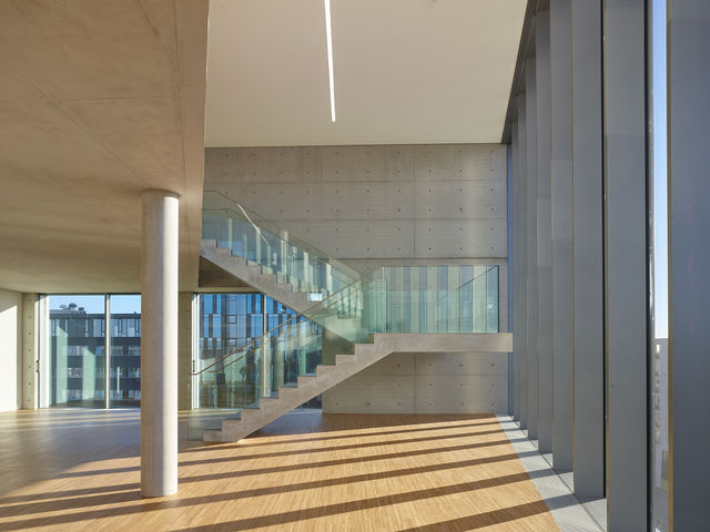 Frei schwebende Betontreppe mit Glasbrüstung vor einer Sichtbetonwand.