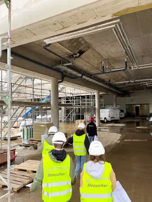 Mädchen und Jungs tragen einen weißen Bauhelm und eine gelbe Warnweste während sie über eine Baustelle laufen
