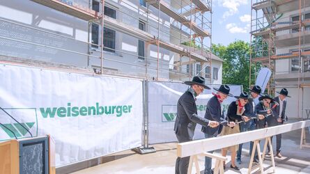 fünf Männer und eine Frau hauen jeweils einen Nagel in einen Holzblock