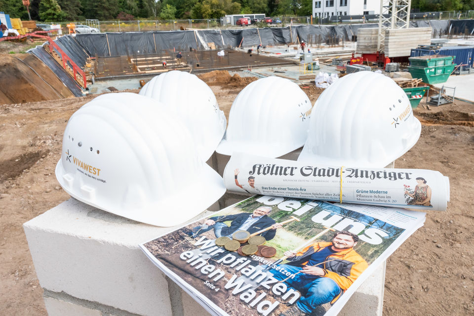 vier weiße Bauhelme und zwei Zeitungen liegen auf einem Stein welcher auf einer Baustelle steht