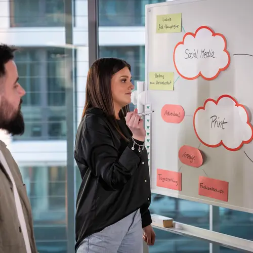 Ein junger Mann und eine junge Frau stehen vor einem Whiteboard und sind auf der Ideensuche 