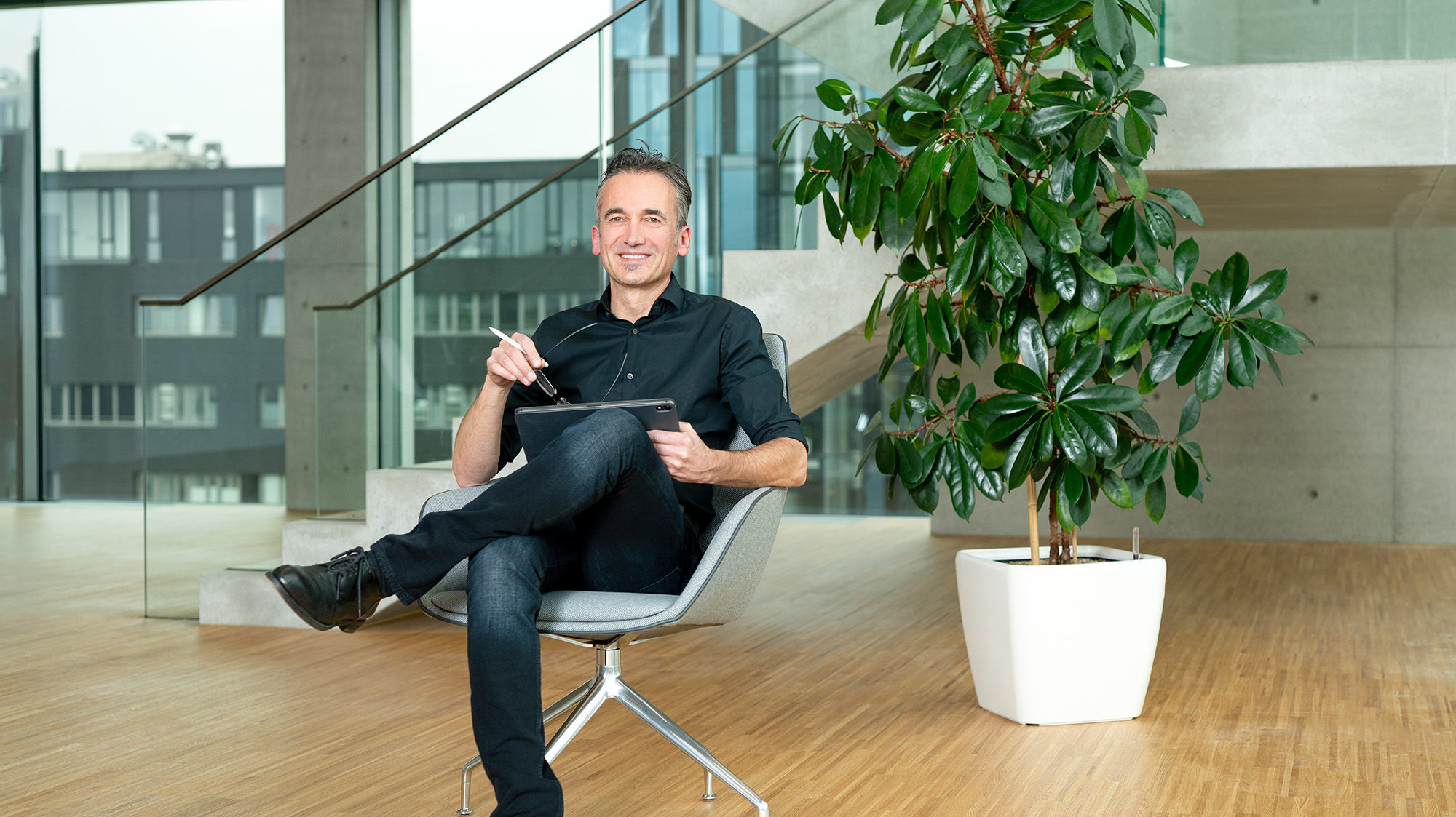  A man sits cross-legged in a chair and looks at the camera smiling