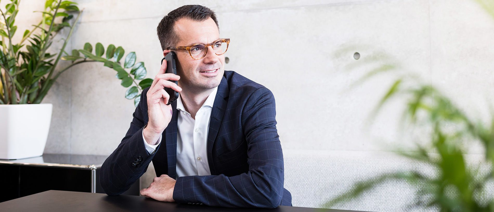 A man with glasses is sitting at a table in front of a houseplant and is talking on the mobile phone