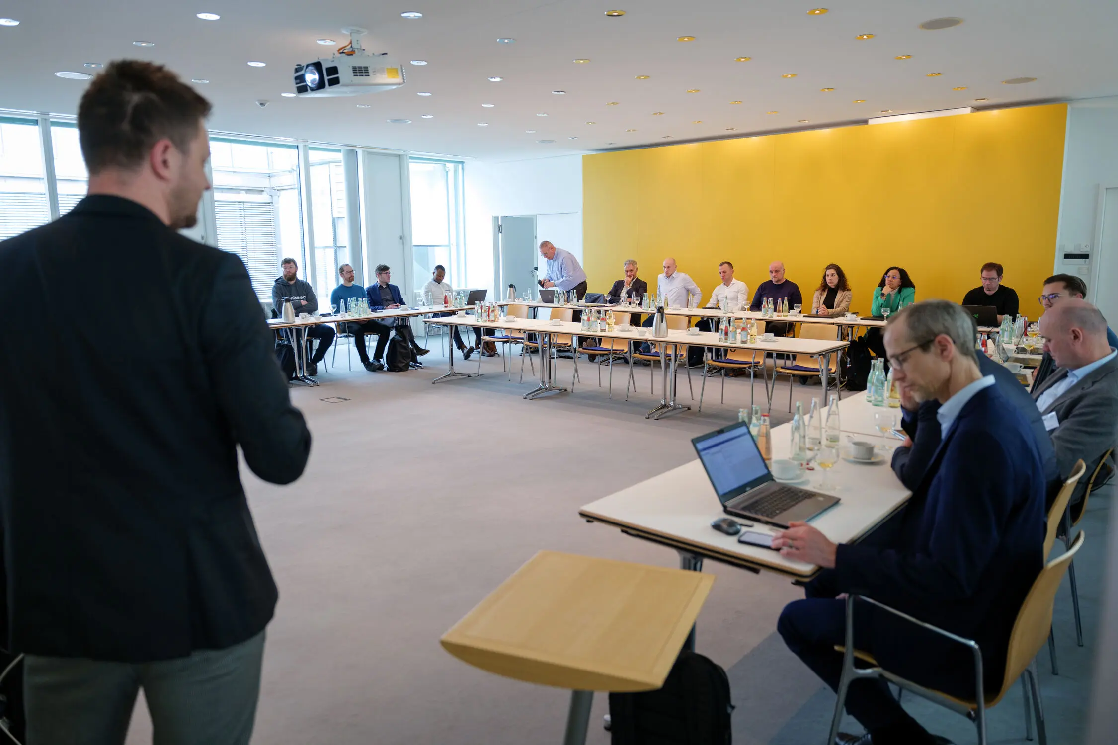 In einem Besprechungsraum wird ein Vortrag von einem Mann gehalten und viele Mitarbeiter hören zu