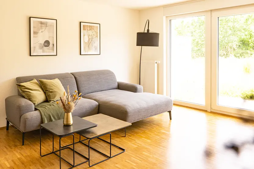 [Translate to English:] Ein graues Sofa mit Liegeflächen und zwei Beistelltischen in einem hellen Raum mit bodentiefen Fenstern und Parkett