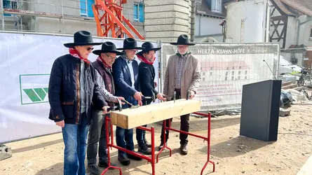 Fünf Männer mit schwarzem Hut stehen auf einer Baustelle und schlagen jeweils einen Nagel in einen Holzblock