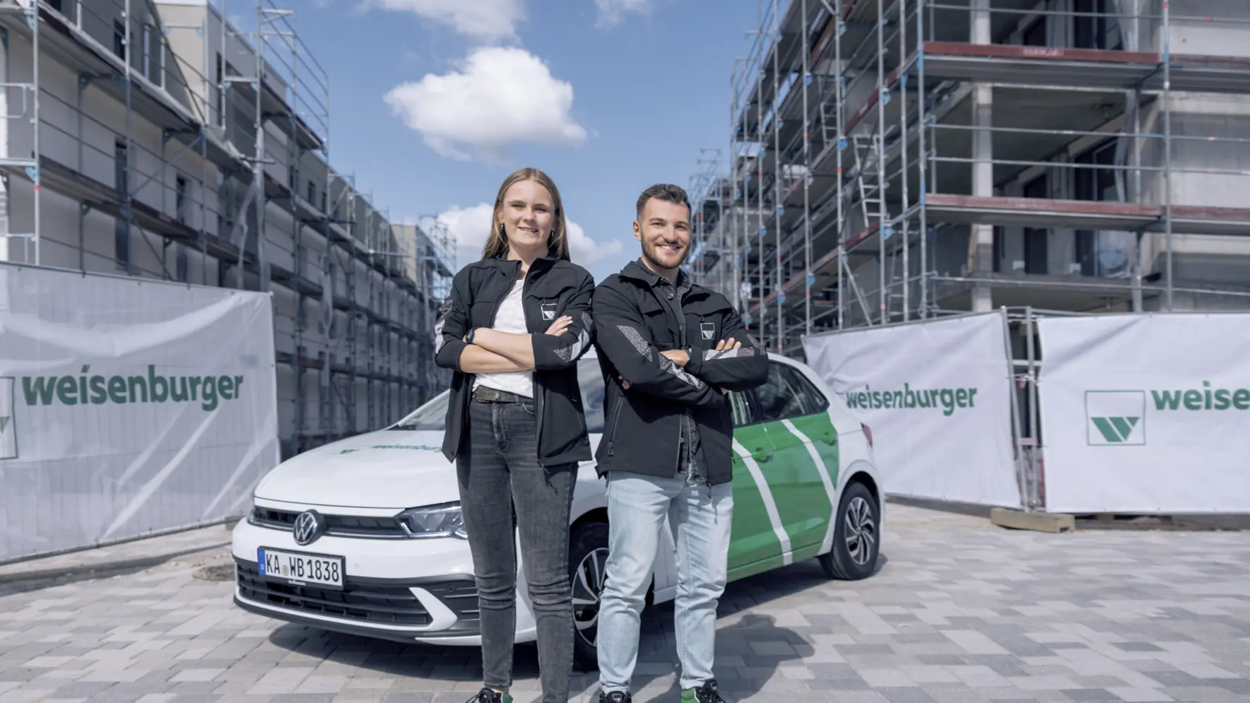 weisenburger-Studierende-Praktikum eine junge Frau und ein junger Mann verschraenken beide die Arme und laecheln, sie stehen vor einem weisenburger Auto auf einen Baustelle