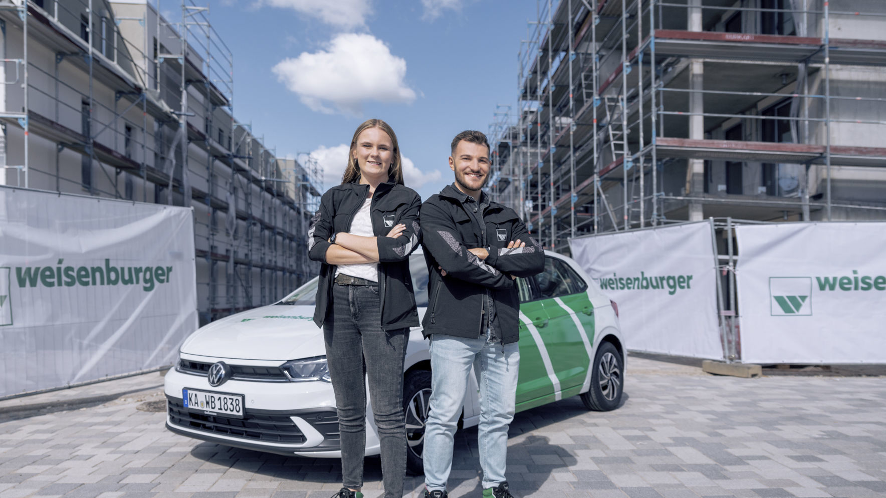 weisenburger-Studierende-Praktikum eine junge Frau und ein junger Mann verschraenken beide die Arme und laecheln, sie stehen vor einem weisenburger Auto auf einen Baustelle