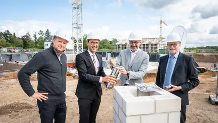 Vier Männer mit Bauhelmen legen auf einer Baustelle symbolisch eine Zeitkapsel in einen Grundstein.