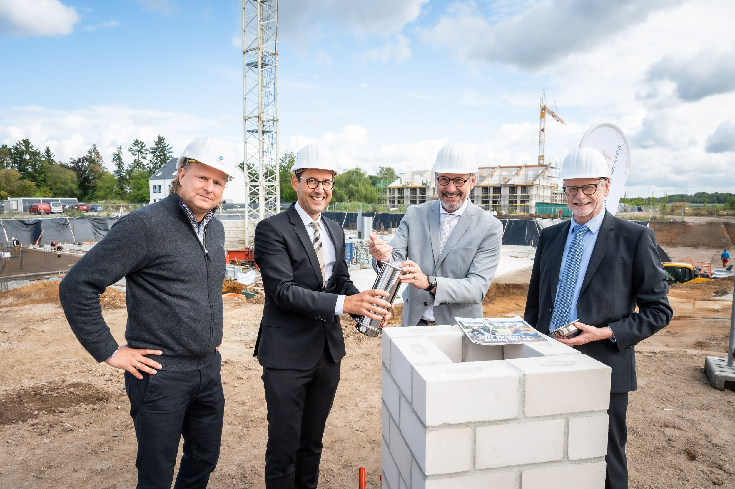 Vier Männer mit Bauhelmen legen auf einer Baustelle symbolisch eine Zeitkapsel in einen Grundstein.