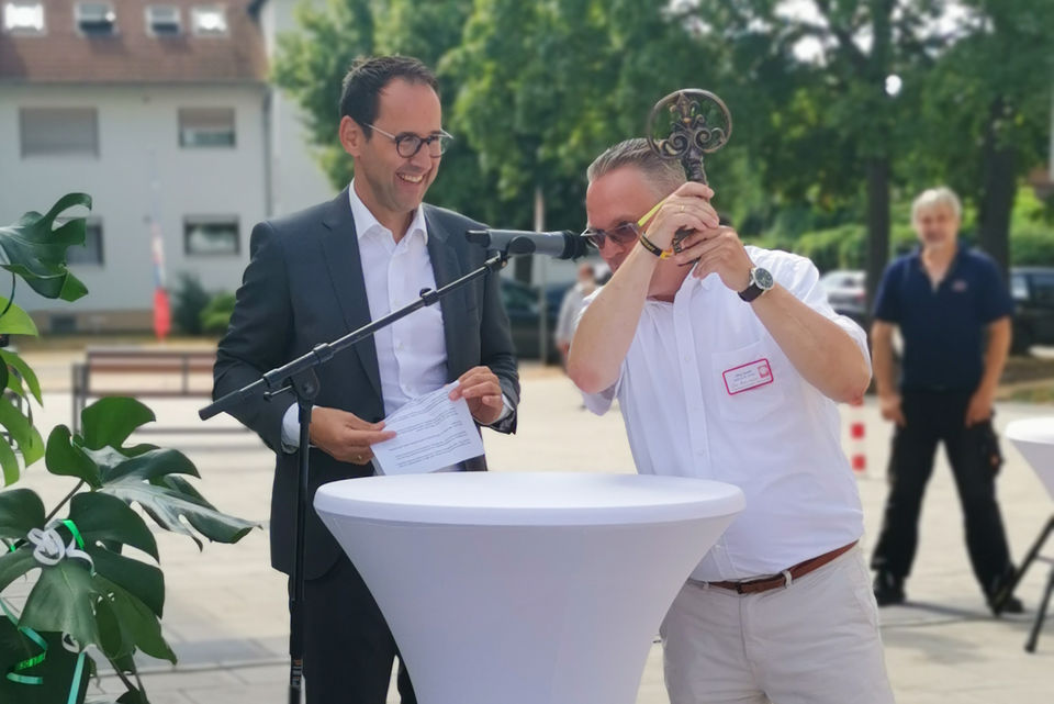 Zwei Männer bei einer Schlüsselübergabe im Freien, wobei einer den symbolischen Schlüssel hochhält und beide fröhlich lachen