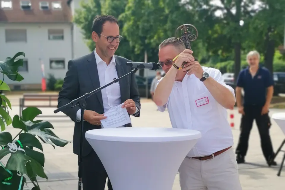 Zwei Männer bei einer Schlüsselübergabe im Freien, wobei einer den symbolischen Schlüssel hochhält und beide fröhlich lachen