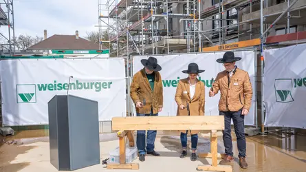 Drei Personen in Handwerkskleidung stehen auf einer Baustelle vor einem Holzbalken mit "weisenburger"-Banner im Hintergrund