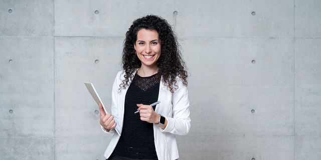 Eine Mitarbeiterin posiert lächelnd vor einer Betonwand, in den Händen hält sie einen Notizblock und einen Kugelschreiber
