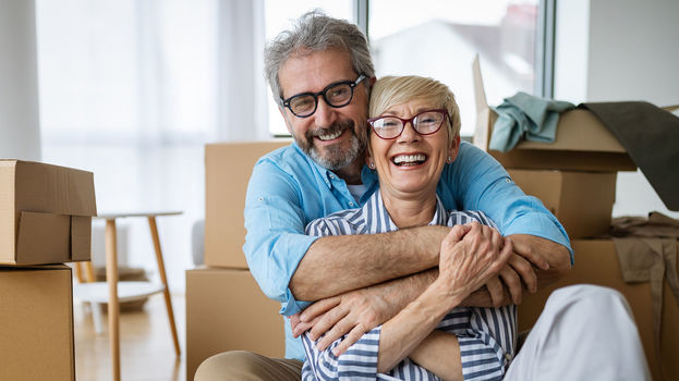 Ein Ehepaar Mitte 50 sitzt lachend inmitten von Umzugskartons.
