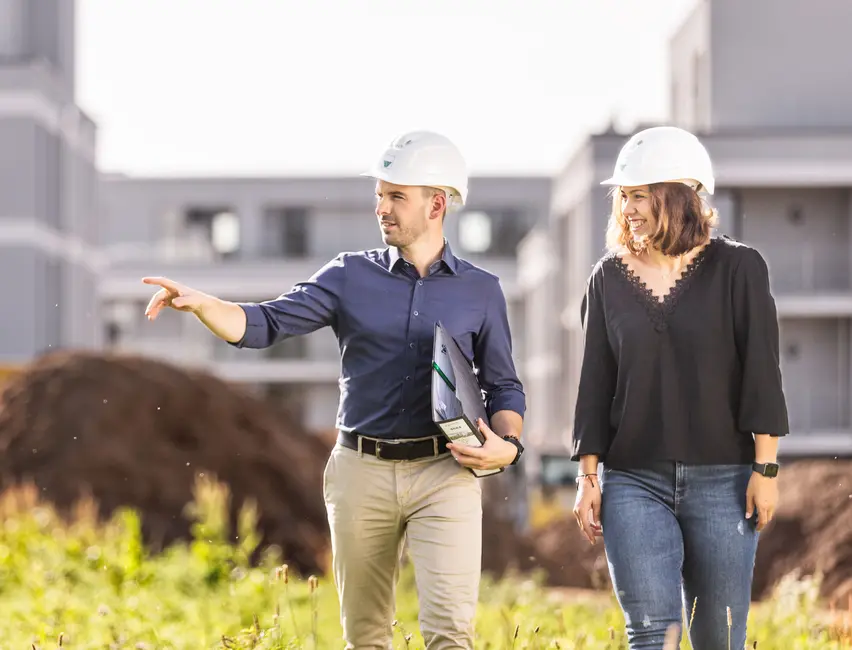 Ein Mann und eine Frau mit Bauhelmen laufen lächelnd über eine Baustelle, der  Mann hält in seiner Hand ein Ordner