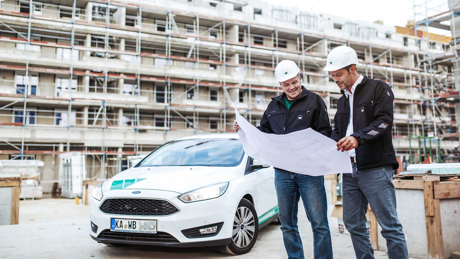 two men are standing in front of a company´s car in front of a construction site and are looking at a construction plan together