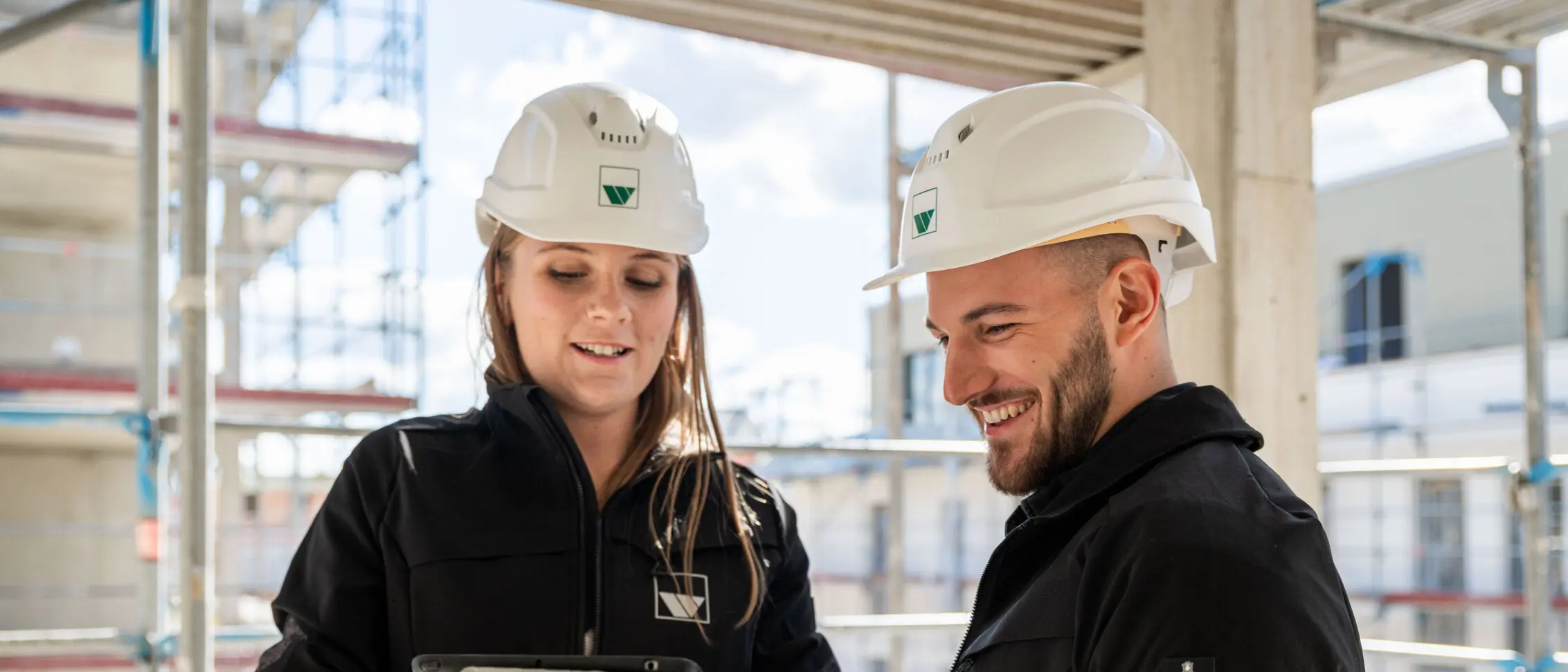 weisenburger-Studium-Jungbauleiter eine junge Frau und ein junger Mann, mit weißen Bauhelmen, stehen auf einer Baustelle und schauen gemeinsam auf ein Tablet